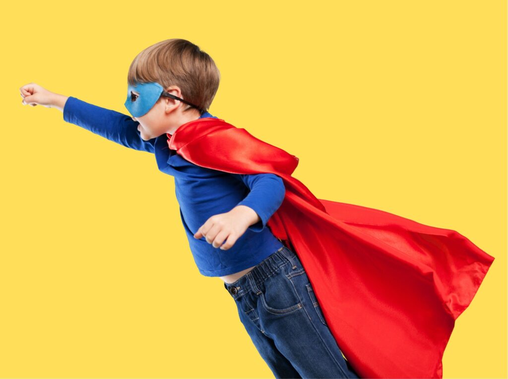 Chłopiec z wyciągniętą w górę pięścią w niebieskiej masce i w czerwonej pelerynie jak Supermana. Szykuje się do lotu. Żółte tło. Dziecko symbolizuje korepetycje w domach dziecka