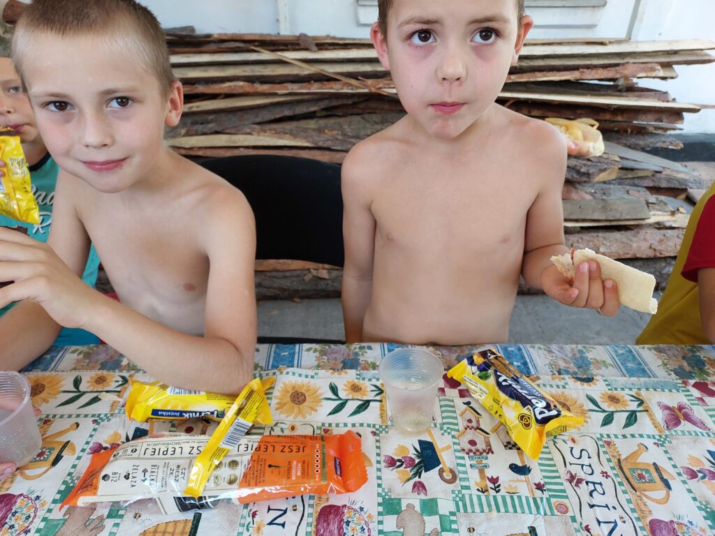Dwóch chłopców bez koszulek siedzi pry stole pokrytym kolorową ceratą. W tle widać stertę desek. Przed chłopcami leżą kabanosy w paczce i paczki pierników. Jeden z chłopców trzyma w ręku zwiniętą tortillę, drugi je piernika.
